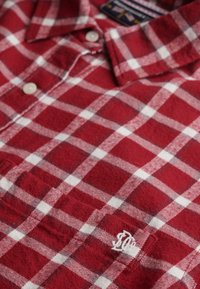 Chemise en flanelle à carreaux rouges et blancs avec un col boutonné, dotée de boutons blancs et d'un logo brodé sur la poche. Texture douce.