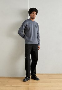 Sivi dukserica s okruglim izrezom i detaljem logotipa na prsima, uparen s crnim ravnim hlačama i crnim cipelama. Pozadina od drvenog poda.