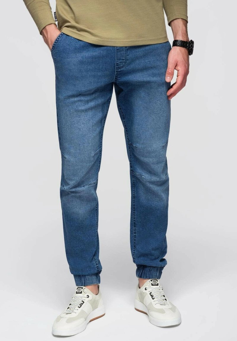 Cómodos pantalones jogger azules con puños elásticos, una textura de denim suave y bolsillos frontales, combinados con zapatillas blancas.