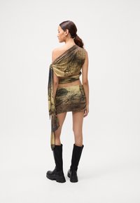 Frau mit einem tiefen Pferdeschwanz, die ein einseitig geschnittenes Crop-Top mit Schriftzug und einen passenden Minirock trägt, dazu schwarze knielange Stiefel, mit dem Rücken zur Kamera gewandt.