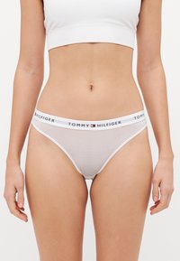 Baltās svītrainās bikini apakšbikses ar zīmola elastīgu jostasvietu, uz kuras ir "TOMMY HILFIGER" logo. Gluda auduma un pieguloša piegriezuma dizains.