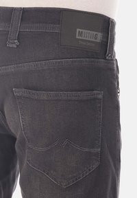 Jeans de mezclilla negra con una superficie texturizada, que cuentan con dos bolsillos traseros y una etiqueta negra rectangular con la inscripción "MUSTANG True Denim" en relieve.