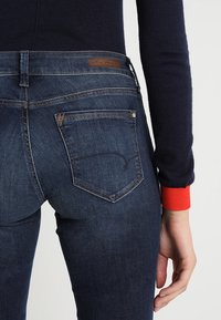 Ciemnoniebieskie dżinsy o pięciokomorowym fasonie, z skórzaną metką na pasku i kontrastującym czerwonym mankietem swetra.