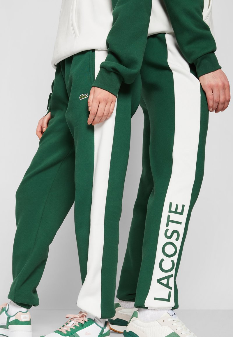 Pantaloni jogger verdi con strisce laterali bianche e testo "LACOSTE". Tessuto morbido, polsini elasticizzati e dettagli con logo. Abbinati a calzature sportive.