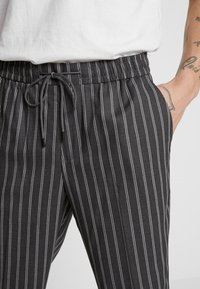 Mustat ja harmaat raidalliset housut, joissa on elastinen vyötärö ja nyöri, varustettuna taskulla ja sileällä, teksturoidulla kankaalla.