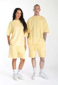 Gule bomulds oversized T-shirts og matchende shorts med en glat overflade. Begge har et logo i hvidt, suppleret med hvide sneakers og sokker.
