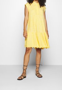 Gul blomsterkjole med høj halsudskæring, korte kappærmer, lagdelt skørt og bølget kant. Kombineret med sorte snøresandaler.