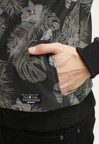 Mano in tasca di un felpa a fantasia floreale scura con l'etichetta del marchio "YOUR TURN" visibile sul lato in basso a sinistra.