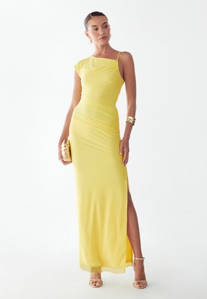 Vrouw in een lange gele een-schouder jurk met een hoge split tot aan de dij, met een geweven clutch in de hand en gouden sandalen en armbanden.