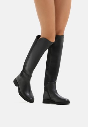 Beine, die schwarze knielange Lederstiefel mit niedrigen Absätzen tragen, stehen vor einem weißen Hintergrund, wobei ein Fuß leicht angehoben ist.