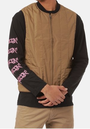 Homme portant une veste matelassée beige sur une chemise noire à manches longues avec un texte graphique rose sur les manches, debout les mains jointes.