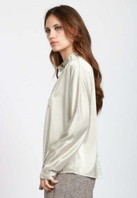 Blusa di seta in beige chiaro, con maniche lunghe, una leggera lucentezza e una vestibilità morbida. Dettagli notevoli sul colletto e bottoni decorativi sulle maniche.