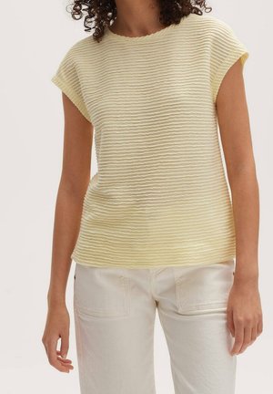 Woman wearing a short-sleeve, light yellow textured top and off-white pants with front patch pockets, standing with relaxed arms.