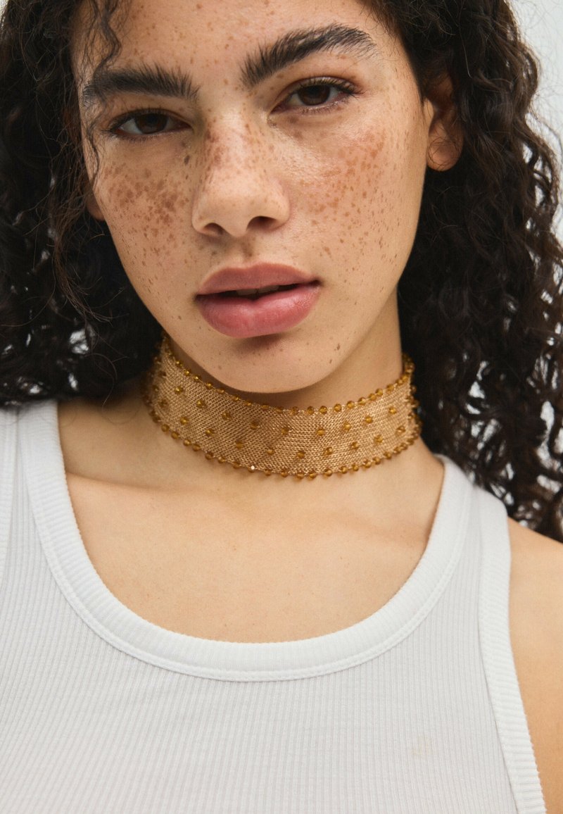 Jeune femme aux cheveux noirs bouclés, avec des taches de rousseur, portant un débardeur blanc et un large collier ras du cou en perles dorées.