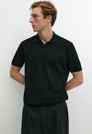 Junger Mann mit hellbraunen Haaren, der ein schwarzes kurzärmeliges geripptes Polohemd und schwarze Hosen trägt, sieht nach links.