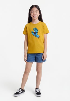 Lachend meisje met lang donker haar, gekleed in een mosterdgele T-shirt met een blauwe schreeuwende hand afbeelding, denim korte broek en zwarte sportschoenen.