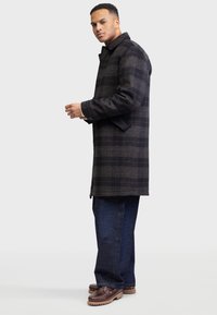 Palton din lână cu carouri în gri închis și negru, cu guler, buzunare laterale și mâneci lungi, purtat cu blugi închiși și pantofi maro.