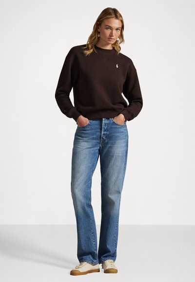 Ciemnobrązowy sweatshirt z długim rękawem i małym logo, w połączeniu z jasnoniebieskimi spodniami prostymi i beżowymi trampkami.