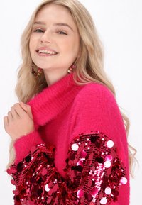 Pulover roz pufoș cu mâneci lungi înfrumusețate cu paiete roșii. Design cu guler înalt, textură moale și detalii colorate vibrante.