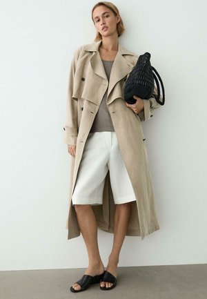 Frau trägt einen beigen Trenchcoat, graues Oberteil, weiße knielange Shorts, schwarze Pantoletten, hält eine schwarze geflochtene Schultertasche.