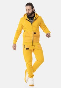 Gelbe Kapuzenjacke und passende Jogginghosen aus weichem Stoff, mit Taschen und kontrastierenden braunen Akzenten. Weiße Sneakers vervollständigen das Outfit.