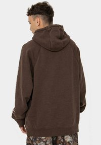 Brązowa bluza z kapturem o teksturowanym wykończeniu, posiadająca kaptur, rękawy raglanowe oraz dolny brzeg. Tył ma minimalistyczne detale i jednolity kolor.