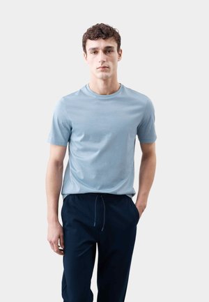 Ung mand med kort krøllet hår iført en lyseblå t-shirt og marineblå bukser med snor, stående med den ene hånd i lommen.