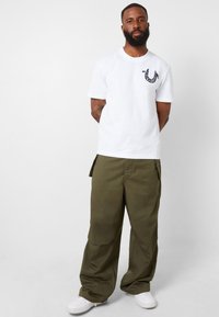 Camiseta de algodón blanca con un pequeño logo azul en el pecho, combinada con pantalones cargo verde olivo y zapatillas blancas.