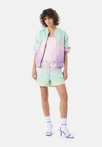 Pastellfarget gradient bomberjakke med lysegrønne og lilla nyanser, kombinert med matchende shorts og lilla detaljerte hæler. Hvite treningssokker.