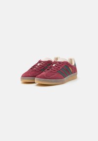 Røde mockaskinnsneakers med grønne striper, kremfargede lisser og fôr. Har en gjennomsiktig gummisåle og "Gazelle" trykt i gull.