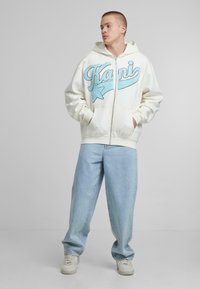 Felpa bianca con zip e testo ricamato blu e stella, abbinata a jeans azzurri larghi e sneakers bianche. Texture del tessuto liscia.
