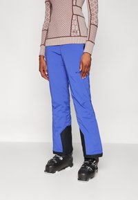 Blauwe skibroek met zwarte accenten, waterdicht materiaal, taps toelopende benen en versterkte boorden. Draagt met zwarte skischoenen.