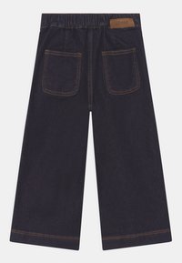 Donkerblauwe wijde denimbroek met elastische tailleband, twee achterzakken, contrasterende oranje stiksels en een klein bruin merklabel.