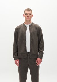 Mørkebrun zip-up bomberjakke med rund halsudskæring, lange ærmer og textured stof, båret over en hvid t-shirt og matchende bukser.