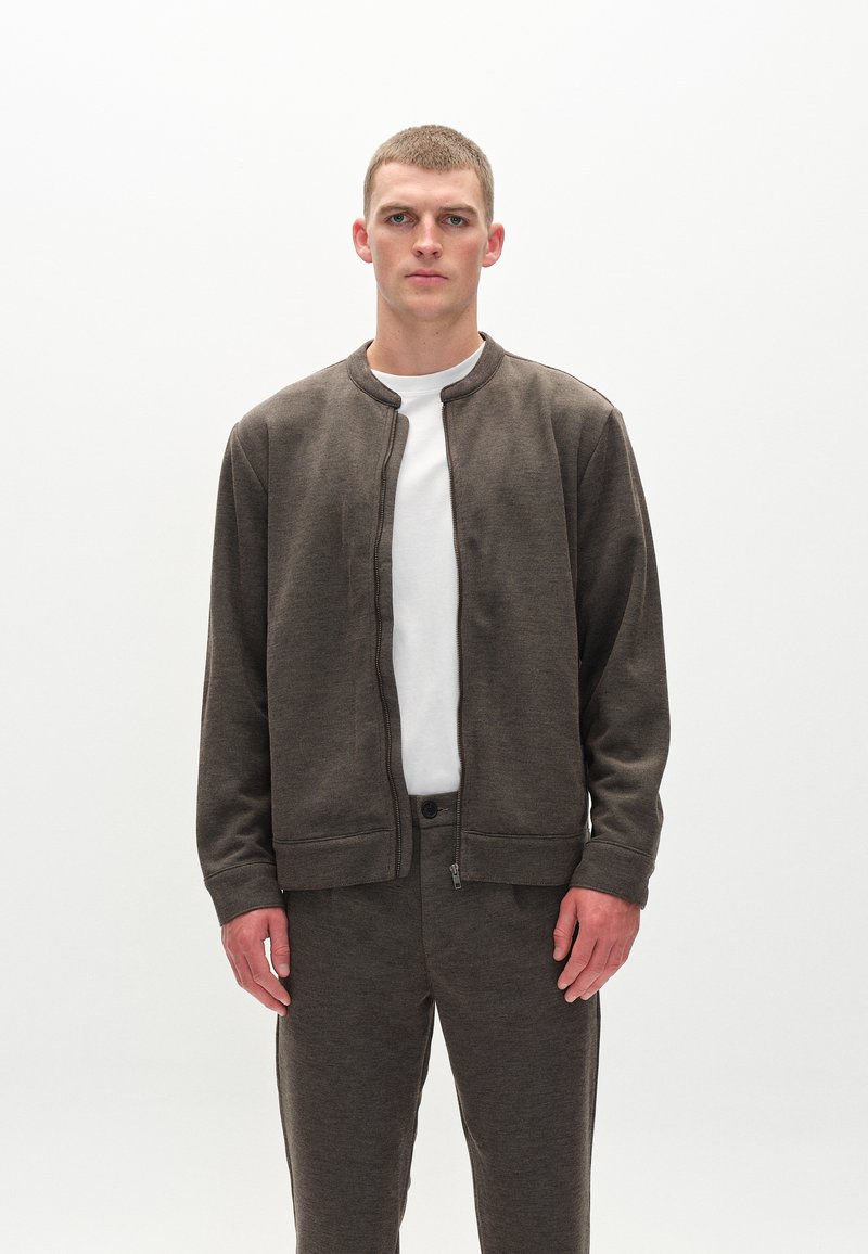 Mørkebrun zip-up bomberjakke med rund halsudskæring, lange ærmer og textured stof, båret over en hvid t-shirt og matchende bukser.
