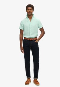 Camicia verde chiaro a maniche corte con bottoni, jeans blu scuro, cintura in pelle marrone e scarpe color sabbia. Il modello sta in piedi con le mani ai fianchi.