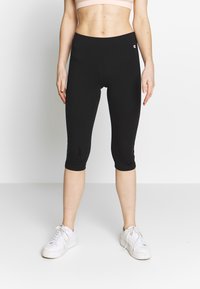 Czarne legginsy capri z elastycznego materiału, z wysokim pasem i małym logo na boku, zestawione z białymi trampkami.