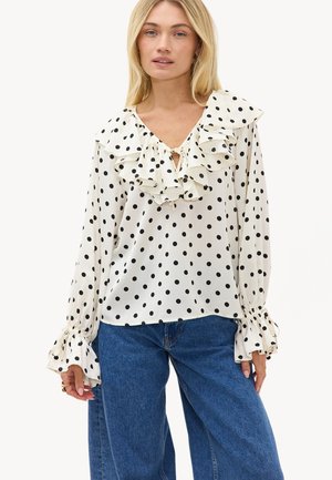 Femme portant un chemisier blanc à manches longues avec des pois noirs, col et poignets volantés, associé à un jean bleu taille haute.