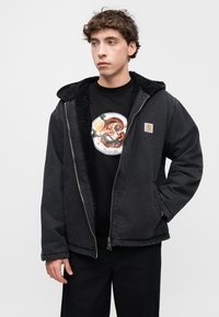 Μαύρο μπουφάν με φερμουάρ και κουκούλα επενδυμένη με γούνα, με ασημένιο φερμουάρ και μικρό λογότυπο. Φορεμένο πάνω από μαύρο μπλουζάκι με κυκλικό σχέδιο.