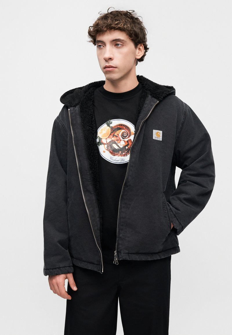 Sort lynlåstrøje med pelsforet hætte, der har en sølvfarvet lynlås og en lille logo-patch. Bærtes over en sort t-shirt med en rund grafik.