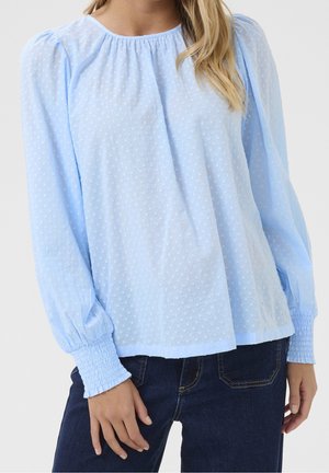 Kvinde iført lyseblå tekstureret langærmet bluse med elastiske manchetter, kombineret med mørkeblå jeans mod en ensfarvet baggrund.