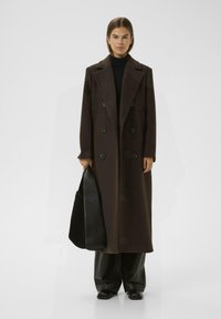 Manteau en laine marron foncé, croisé avec de grands boutons noirs, revers crantés, poches latérales et une coupe ample. Le mannequin tient un sac noir.