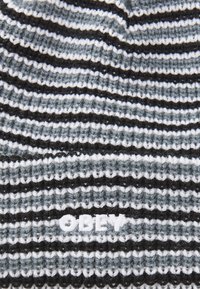 Kootud beanie vahelduvate mustade, valgete ja hallide triipudega. Ees on valge "OBEY" logo tikandiga. Tekstureeritud kangas.