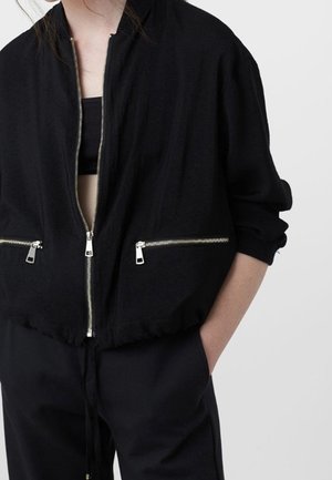 Mujer con chaqueta negra con cremallera plateada sobre una camiseta negra, con las manos en los bolsillos de pantalones negros a juego, sobre un fondo liso.