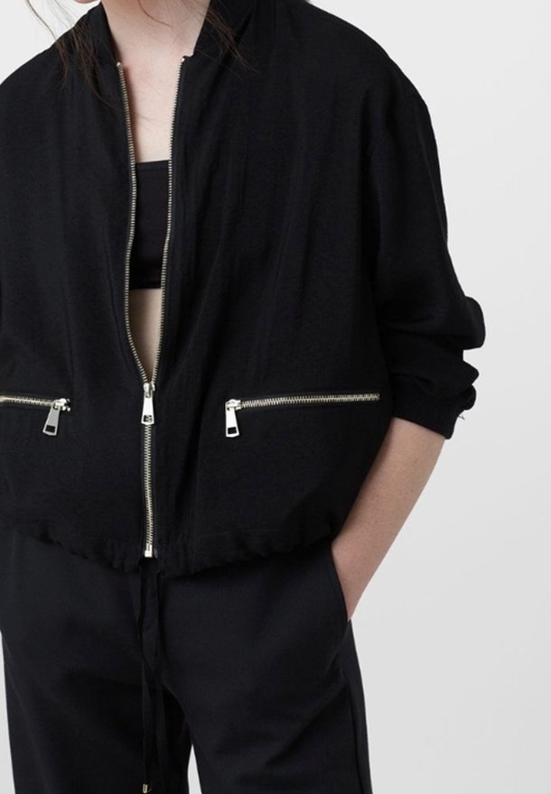 Femme portant une veste noire zippée avec des fermetures éclair argentées par-dessus un haut noir, les mains dans les poches assorties du pantalon noir, sur un fond uni.