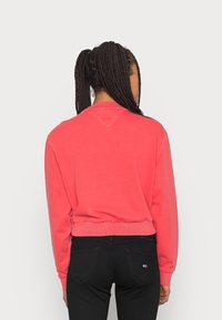 Sweatshirt rosso corto con maniche lunghe, orlo a coste e dettaglio di cucitura a forma di V sul retro, abbinato a jeans neri.