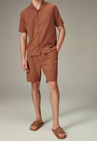 Camicia corta a maniche corte marrone con texture e pantaloni corti abbinati, dotata di una singola tasca sul petto. Indossata con sandali aperti marroni. Design semplice e rilassato.