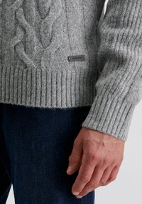 Grau gestrickter Pullover mit Kabelmuster und geripptem Saum, mit einem kleinen Markenetikett an der Seite. Kombiniert mit dunkelblauen Jeans.