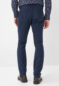 Marinblaue Slim-Fit-Hosen mit glatter Textur, fünf Taschen und einem subtilen Stretch; kombiniert mit braunen Lederschuhen mit minimalen Details.