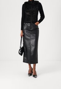 Kvinde iført sort faux pelsvest, ribbet langærmet top, højtaljet lædermidi-nederdel, sorte høje hæle, holder en sort håndtaske.